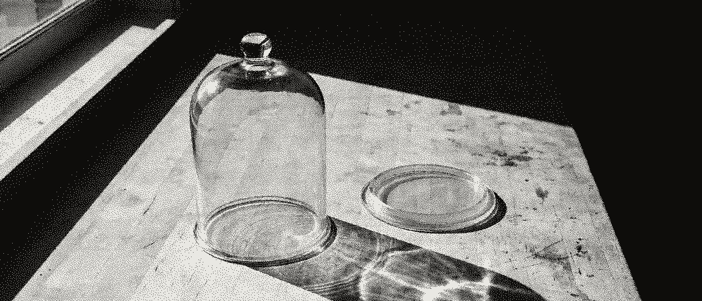 A glass bell jar standing on a pale bench beside its empty base plate, its shadow thrown long across the wood in hard sun.