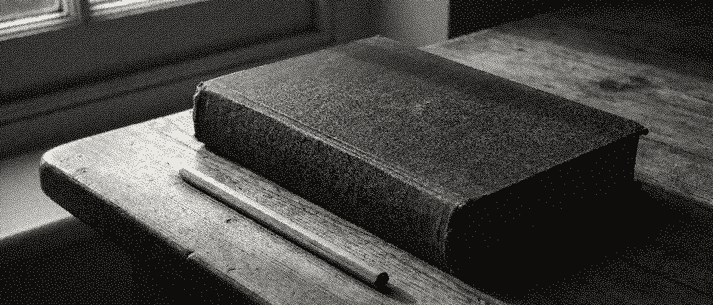 A closed clothbound book without lettering and a pencil on a worn wooden table by a window.
