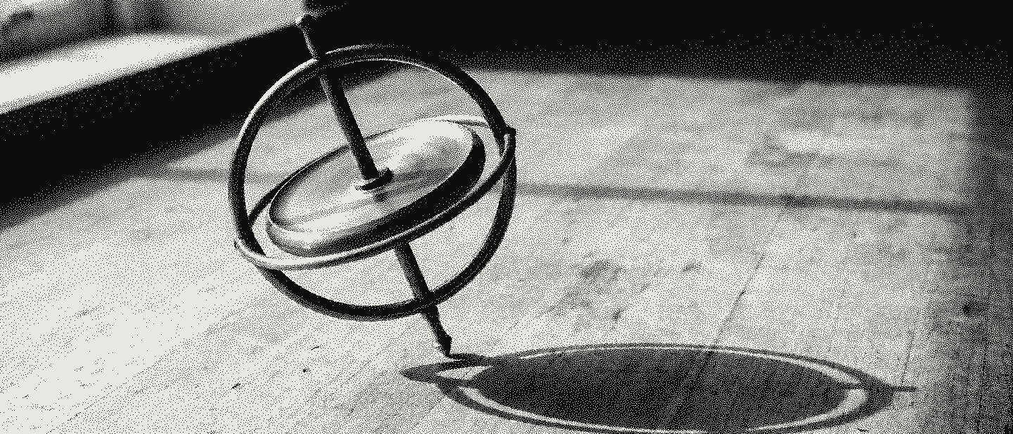 A brass gyroscope balanced at a tilt on the tip of its axle on a pale bench, its shadow an ellipse.