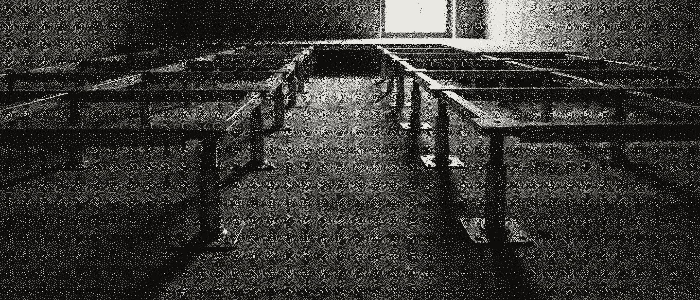 Rows of steel floor pedestals and stringers on a bare concrete slab, an aisle leading to a tall window.